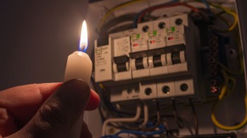 A hand holding a lit candle in front of an electrical circuit breaker box.