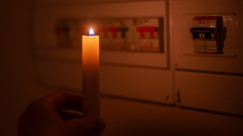 A hand holds a lit candle near an electrical panel in a dimly lit room.