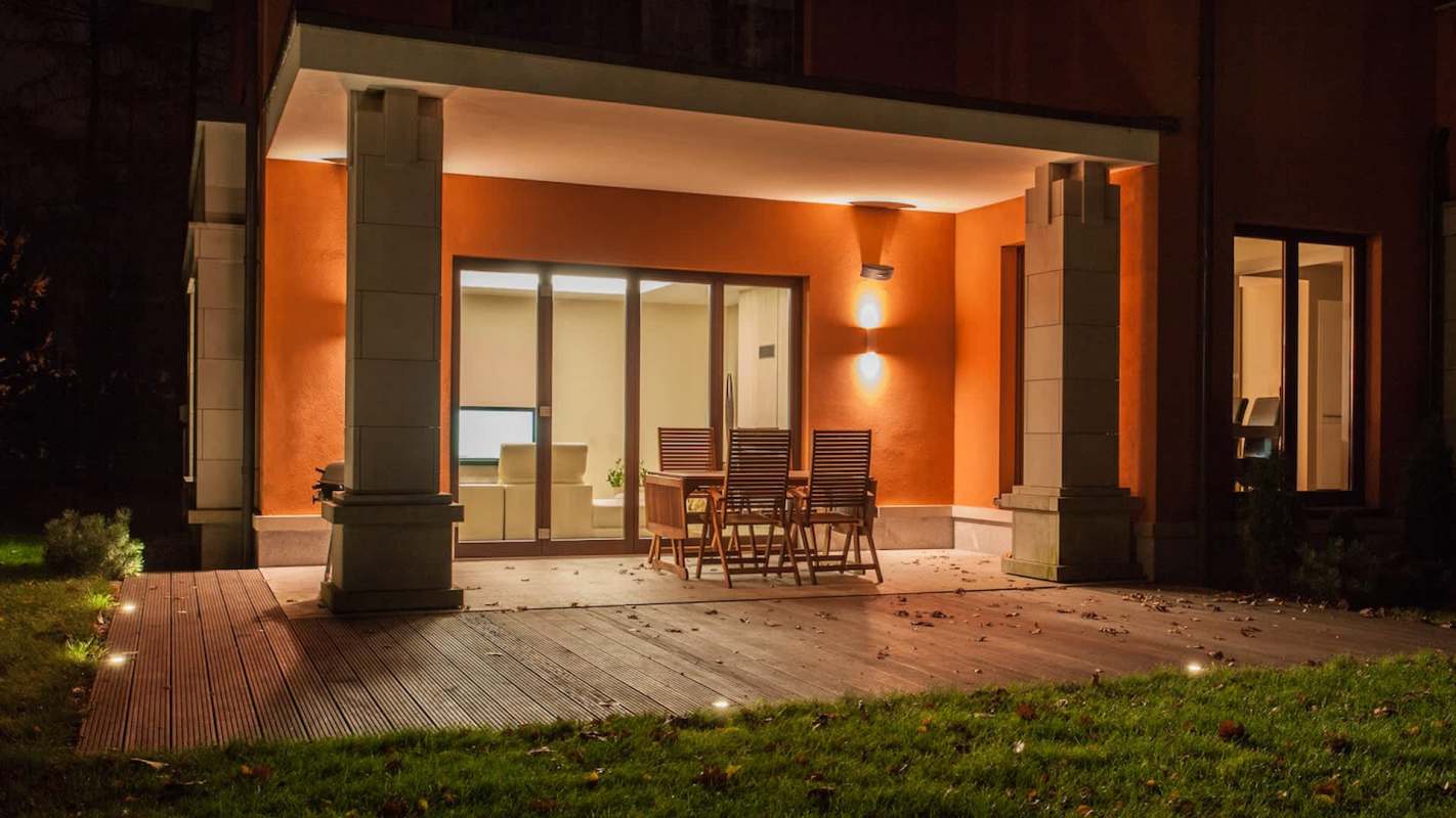 A patio with wooden furniture and a view of a well-lit interior through large glass doors.