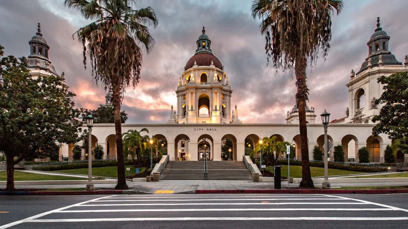 A city hall building with a dome and arched entrance, framed by palm trees and a cloudy sky.