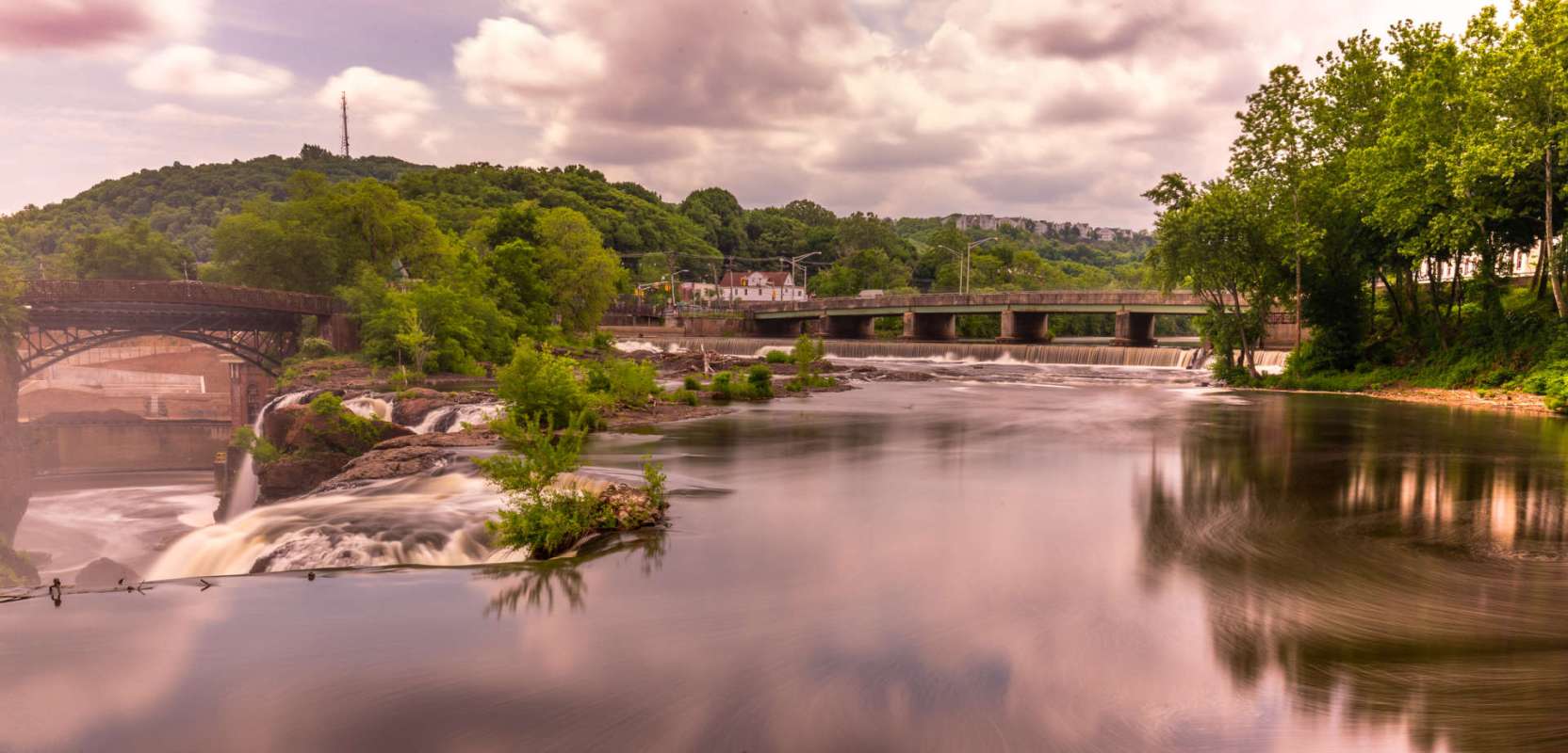 A river flowing past a bridge with trees and a small waterfall in the background under a cloudy sky.