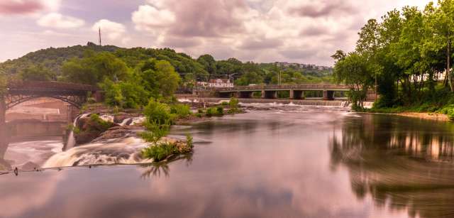 A river flowing past a bridge with trees and a small waterfall in the background under a cloudy sky.