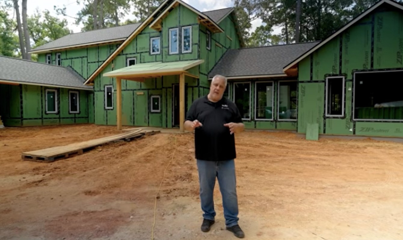 A man stands in front of a partially constructed green house with wooden framing.