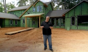 A man stands in front of a partially constructed green house with wooden framing.