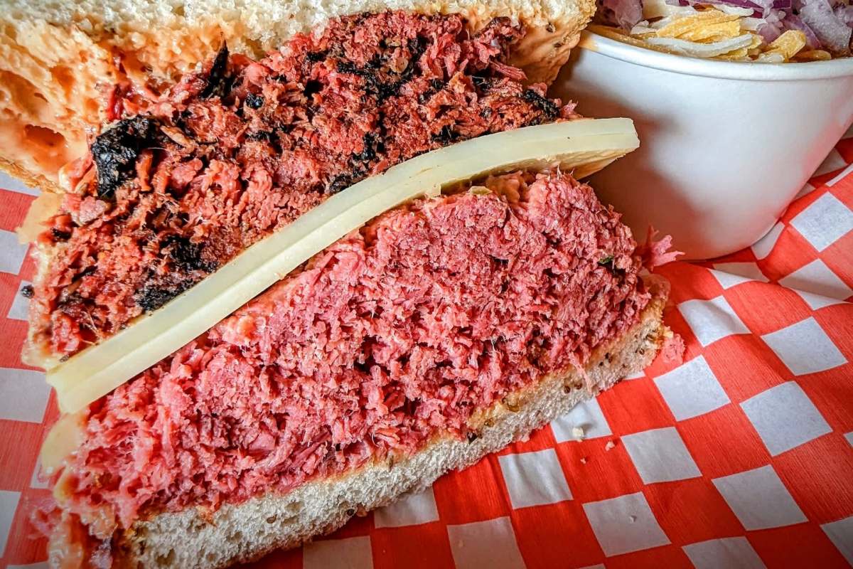 A sandwich with layers of pastrami, corned beef, and a slice of cheese, served with a side of chopped vegetables.