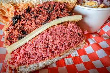 A sandwich with layers of pastrami, corned beef, and a slice of cheese, served with a side of chopped vegetables.