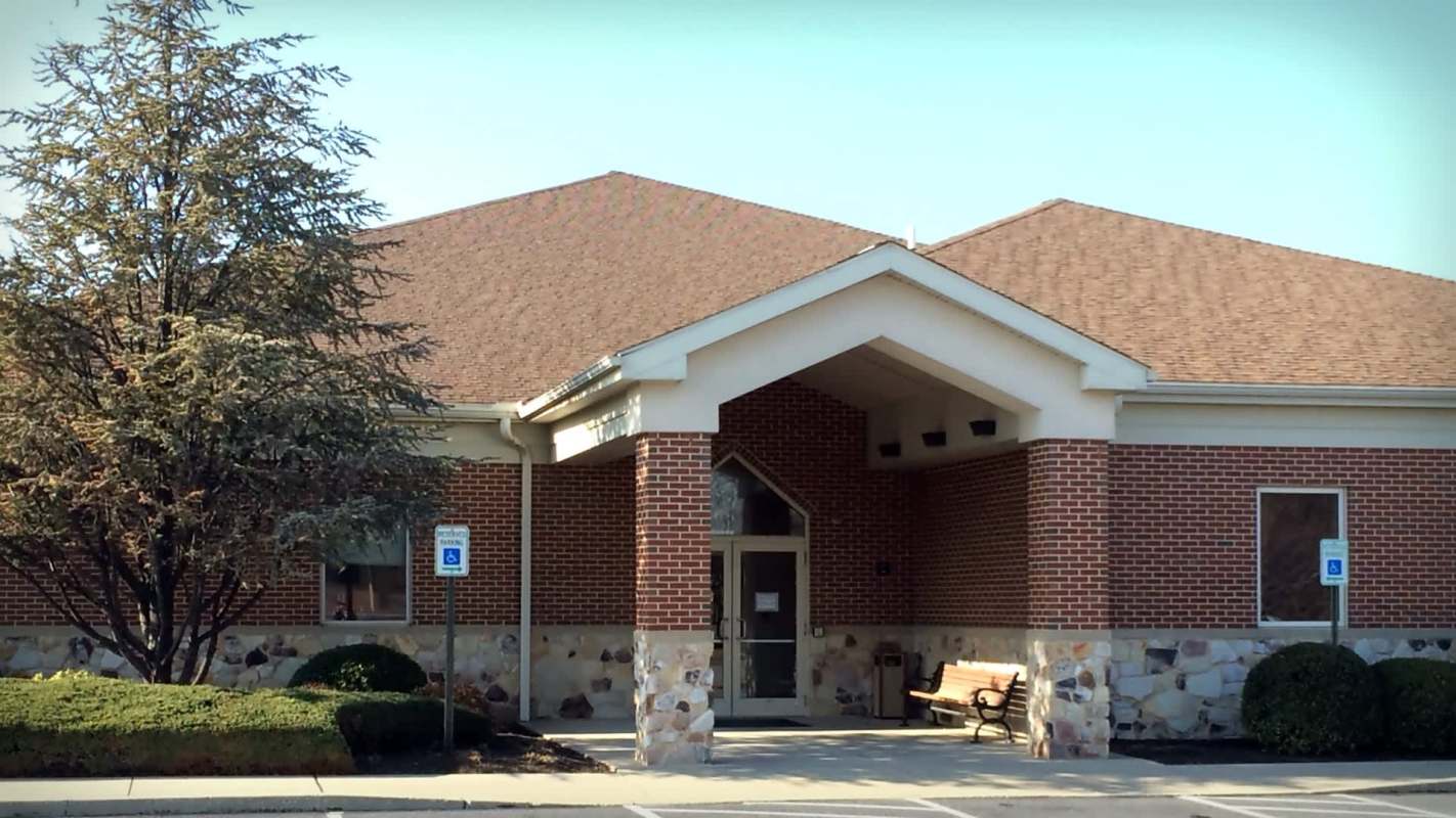 A brick building with a sloped roof and a bench outside, surrounded by shrubs and a parking area.