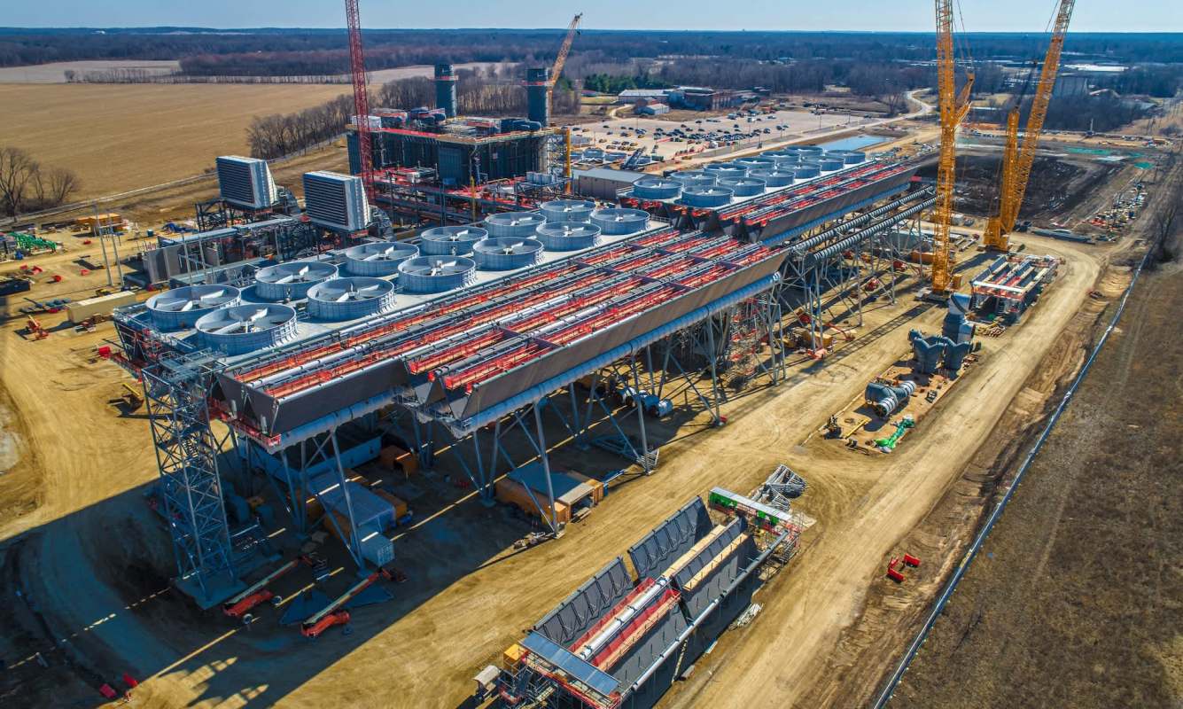 A large industrial facility under construction with cooling towers and cranes in a partially developed area.
