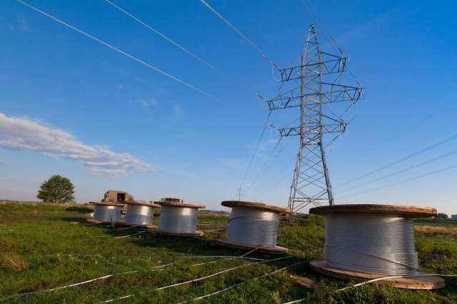 A high-voltage power line tower with large spools of wire in a grassy field under a clear blue sky.