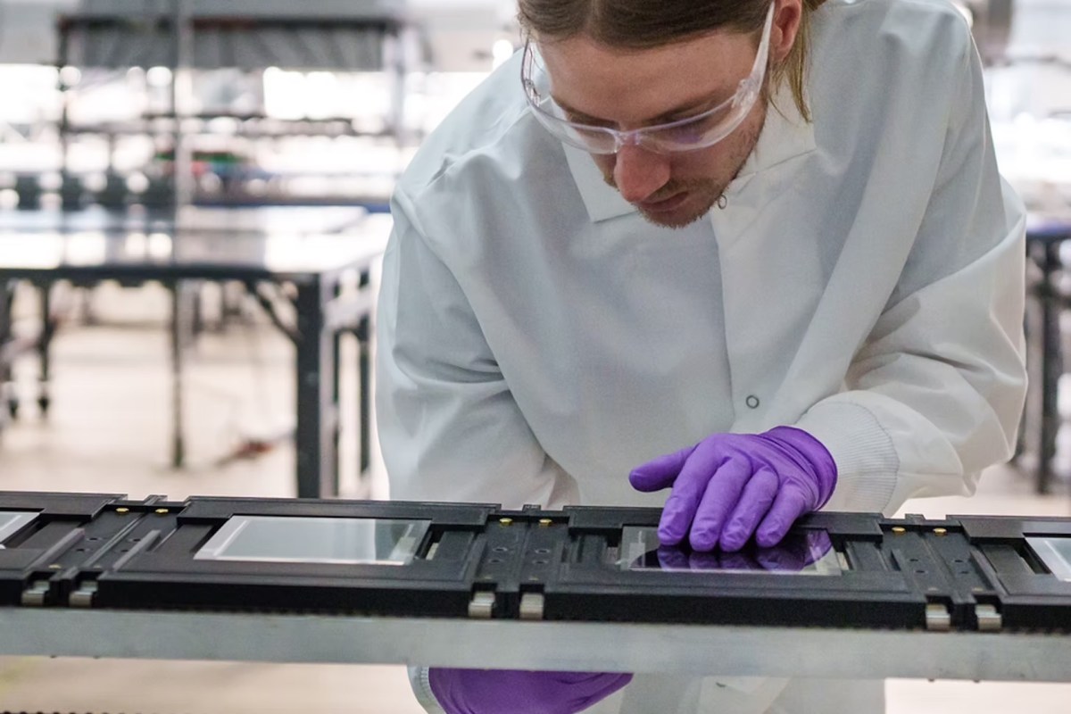 A person in a lab coat and gloves works on a device with glass panels in a laboratory setting.