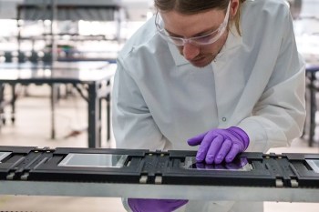 A person in a lab coat and gloves works on a device with glass panels in a laboratory setting.