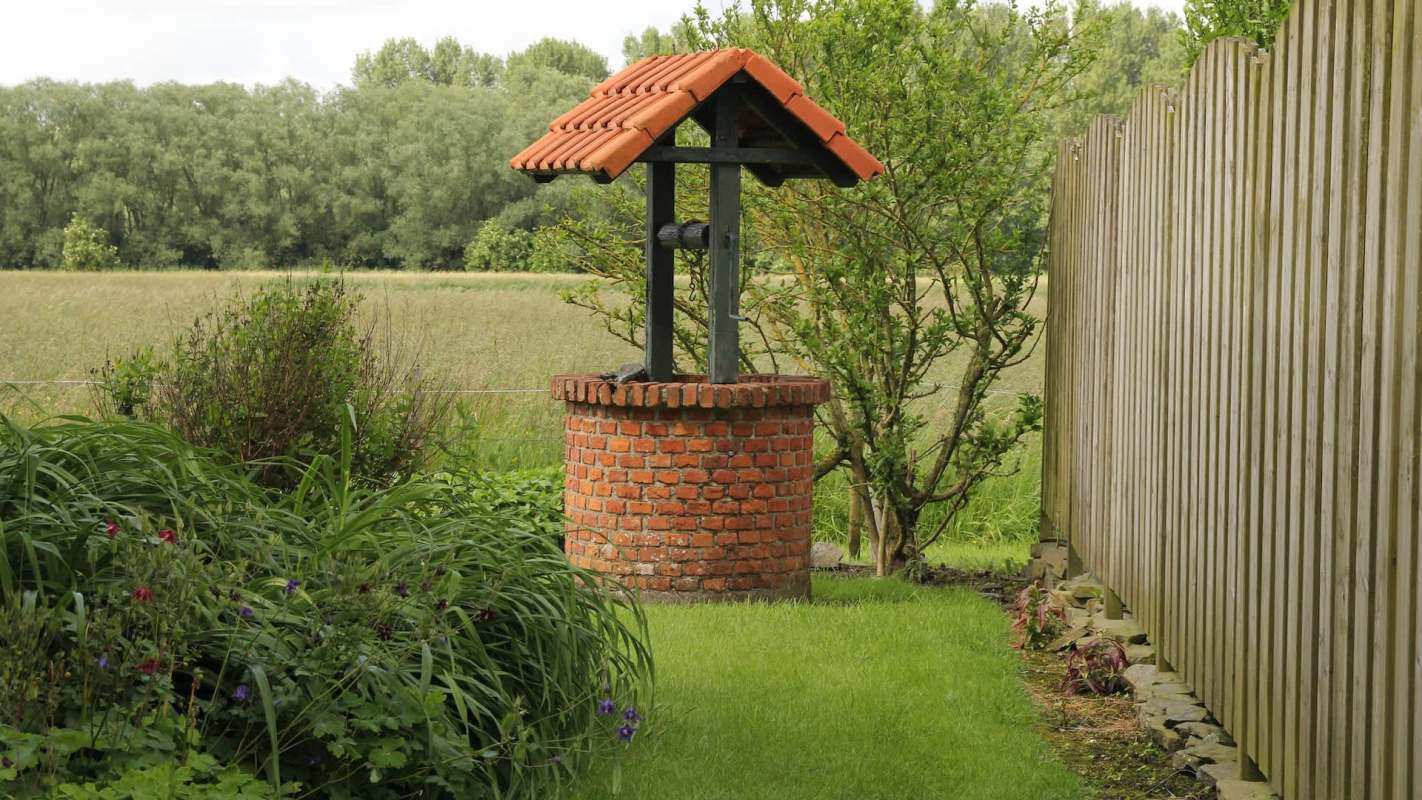 A brick well with a tiled roof surrounded by grass and plants, near a wooden fence and open field.
