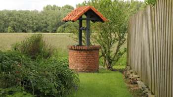 A brick well with a tiled roof surrounded by grass and plants, near a wooden fence and open field.