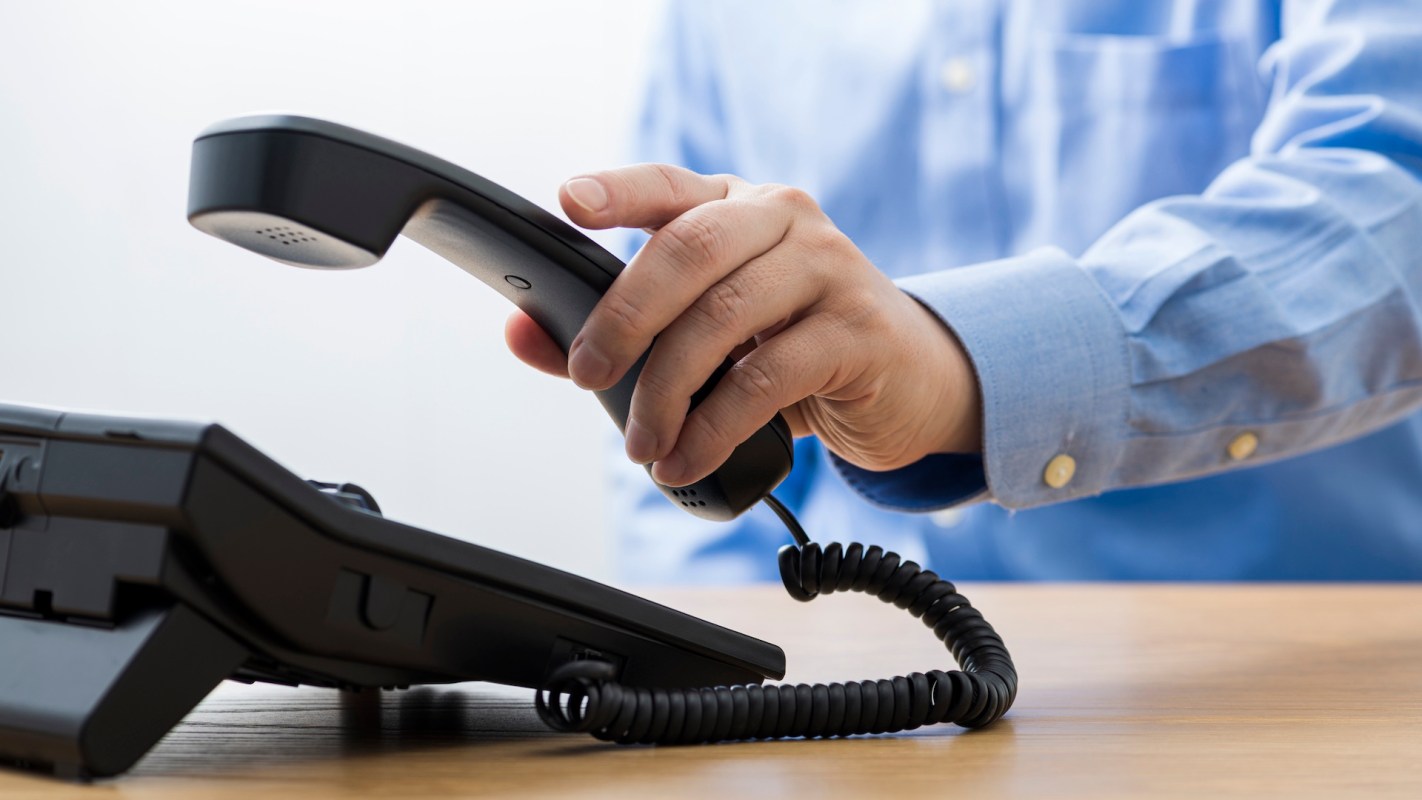 A hand lifts a phone receiver from a black desk telephone.