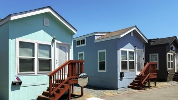 Three small mobile homes with different colors and wooden steps, set against a clear blue sky.