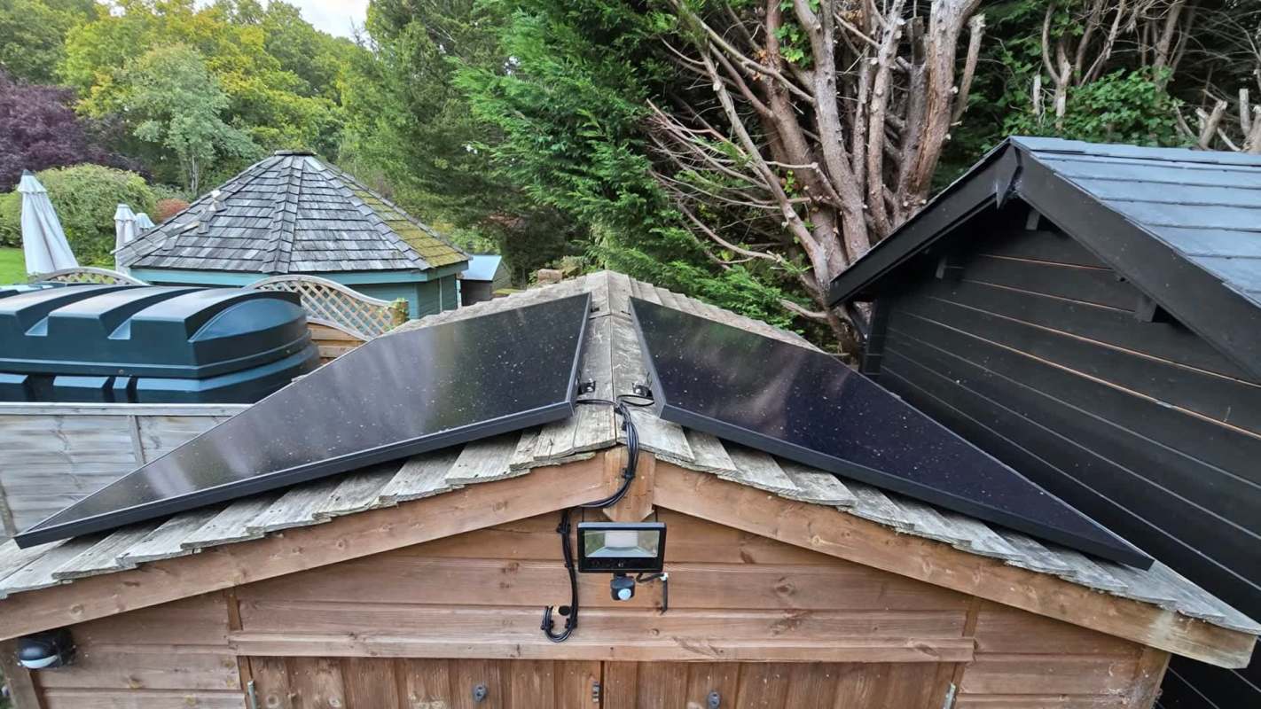 Solar panels installed on a wooden shed roof with greenery and additional buildings in the background.