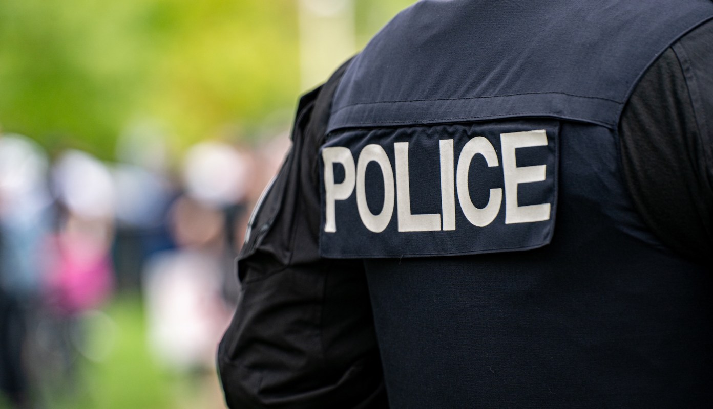 A police officer wearing a uniform with "POLICE" printed on the back stands in a crowd.