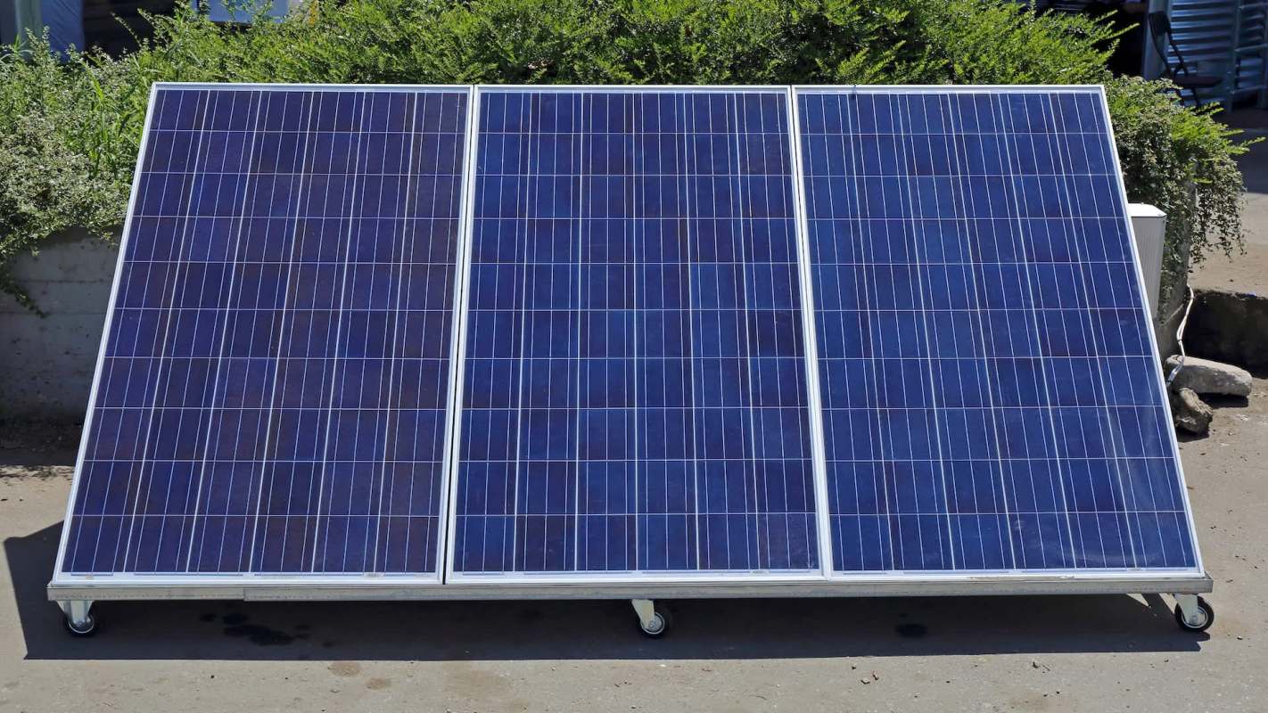 Three solar panels positioned on a wheeled frame with surrounding greenery in the background.
