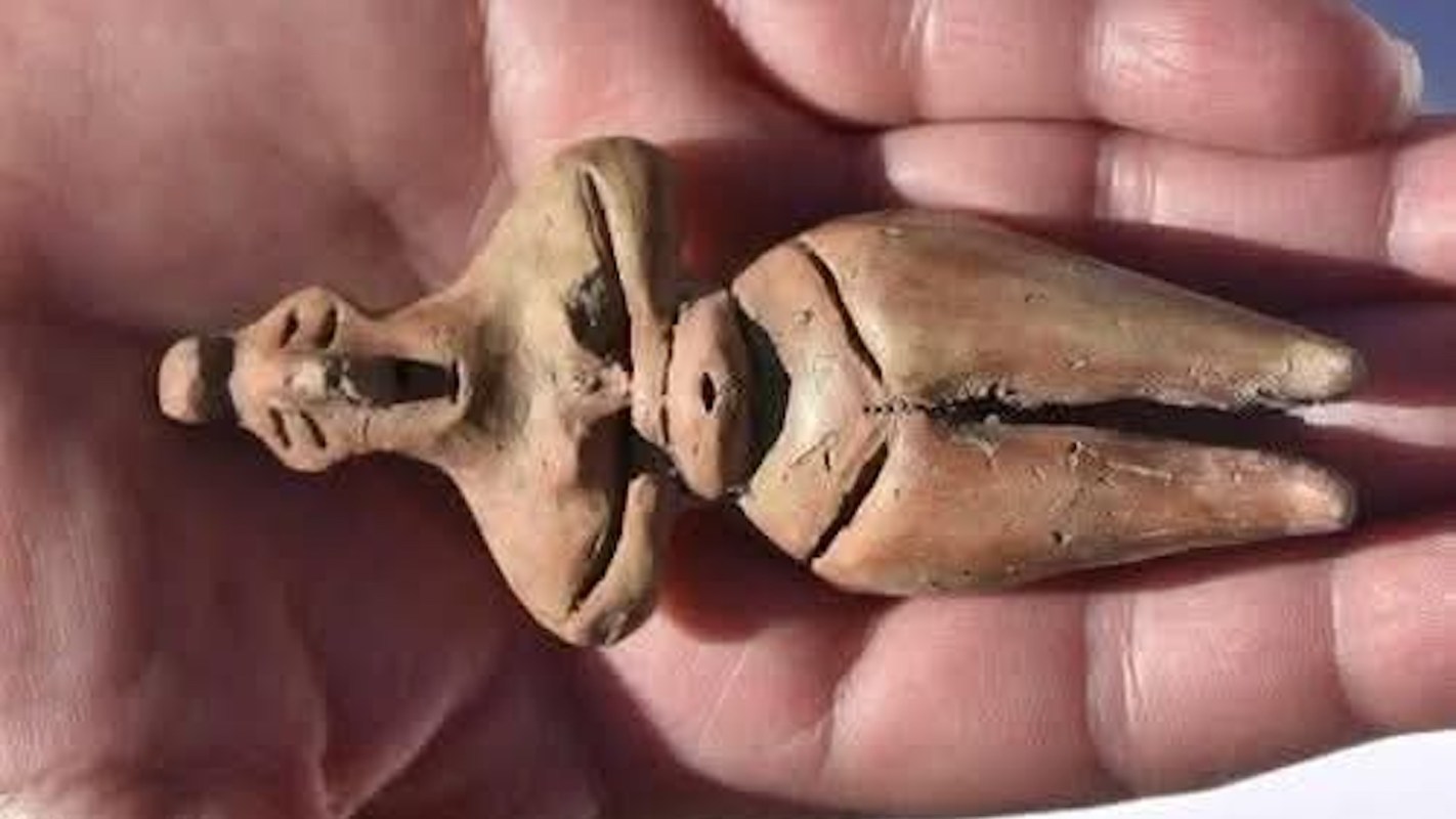 A hand holds a small, ancient clay figurine displaying a humanoid shape with distinct features.