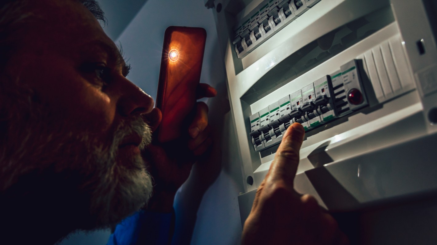 A person examines a circuit breaker panel while holding a phone with a light.