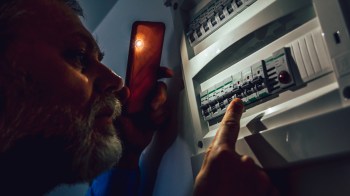A person examines a circuit breaker panel while holding a phone with a light.