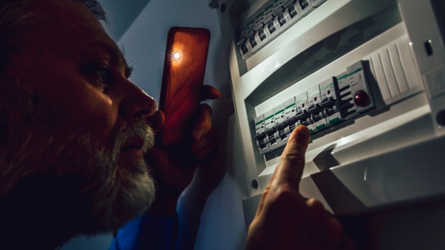 A person examines a circuit breaker panel while holding a phone with a light.