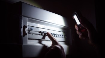 A hand points at a circuit breaker while holding a flashlight in a darkened room.
