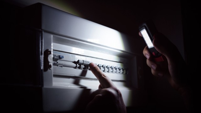 A hand points at a circuit breaker while holding a flashlight in a darkened room.