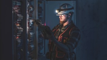 A man in a hard hat and reflective vest works on electrical equipment in a dimly lit environment.