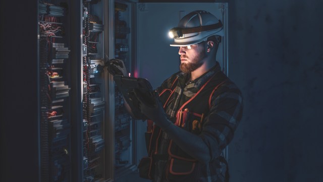 A man in a hard hat and reflective vest works on electrical equipment in a dimly lit environment.