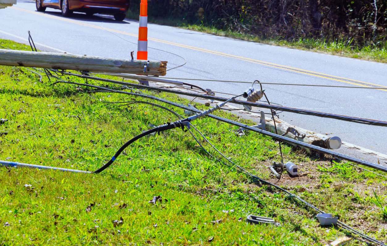 A downed utility pole with tangled wires on the grass beside a road.