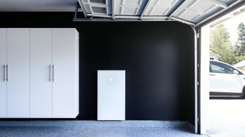 A garage featuring white cabinets, a Tesla Powerwall, and a partially open door revealing a car outside.