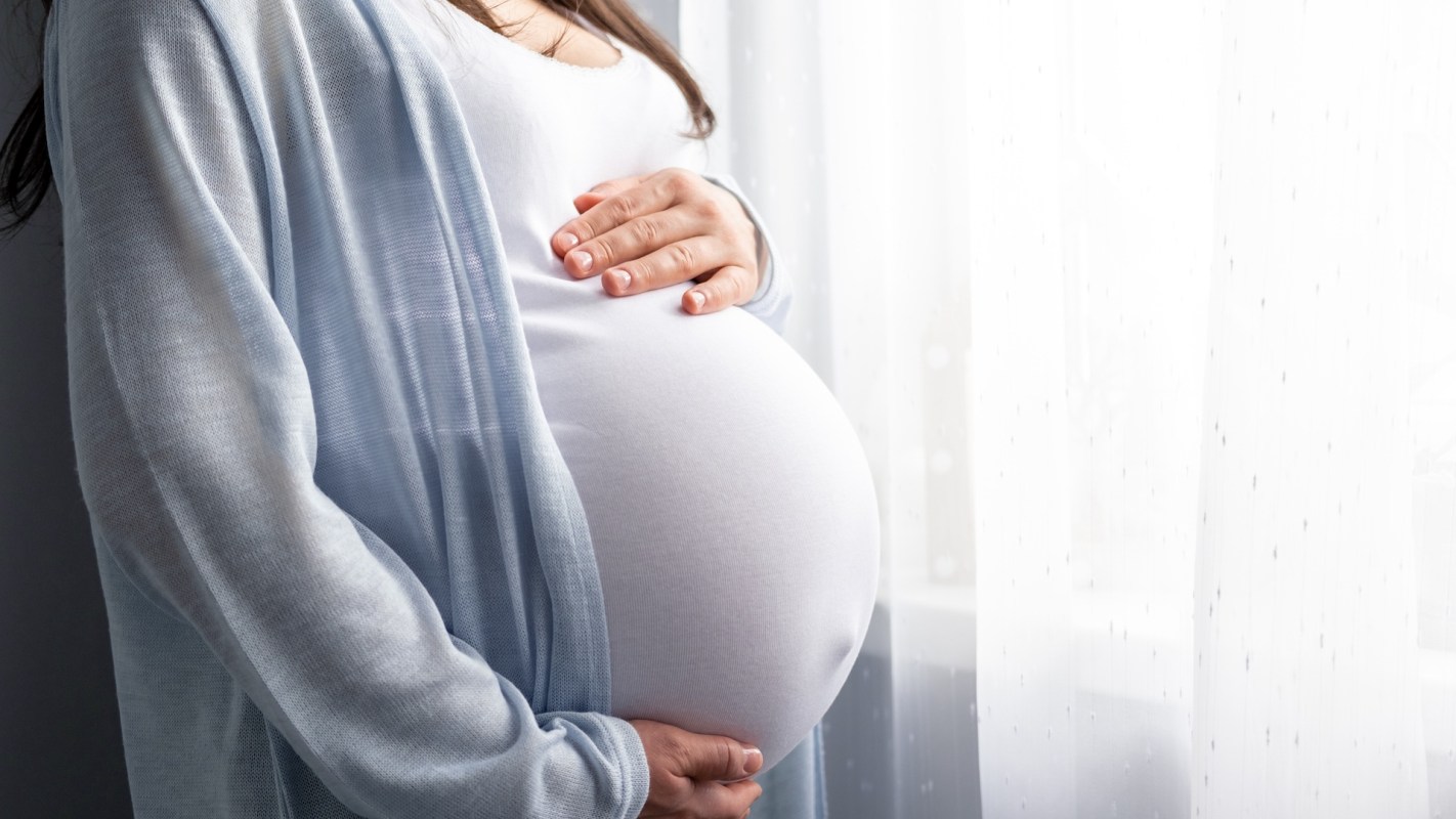 A person holds their pregnant belly in front of a window with sheer curtains.