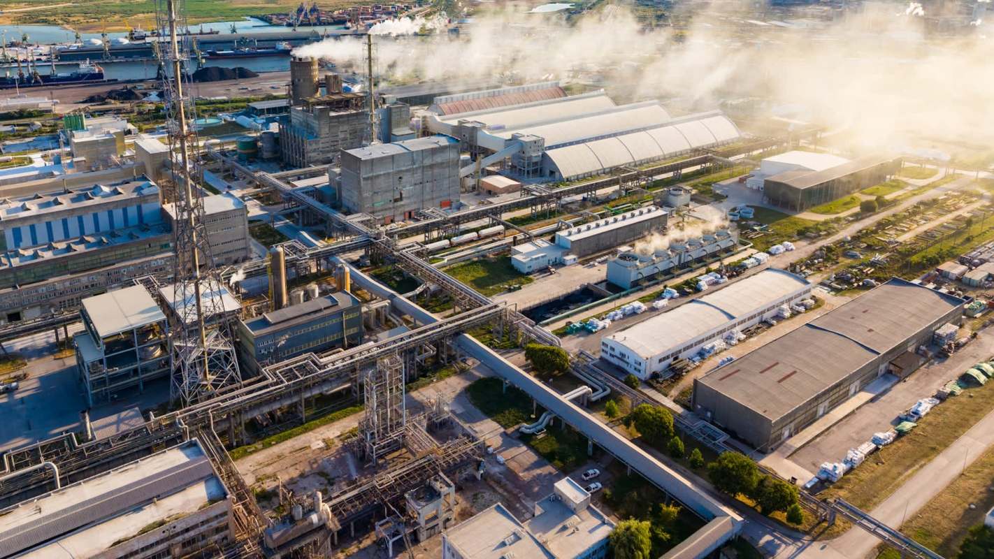 An industrial complex with large buildings, pipes, and smoke stacks, surrounded by warehouses and cargo ships in a harbor.