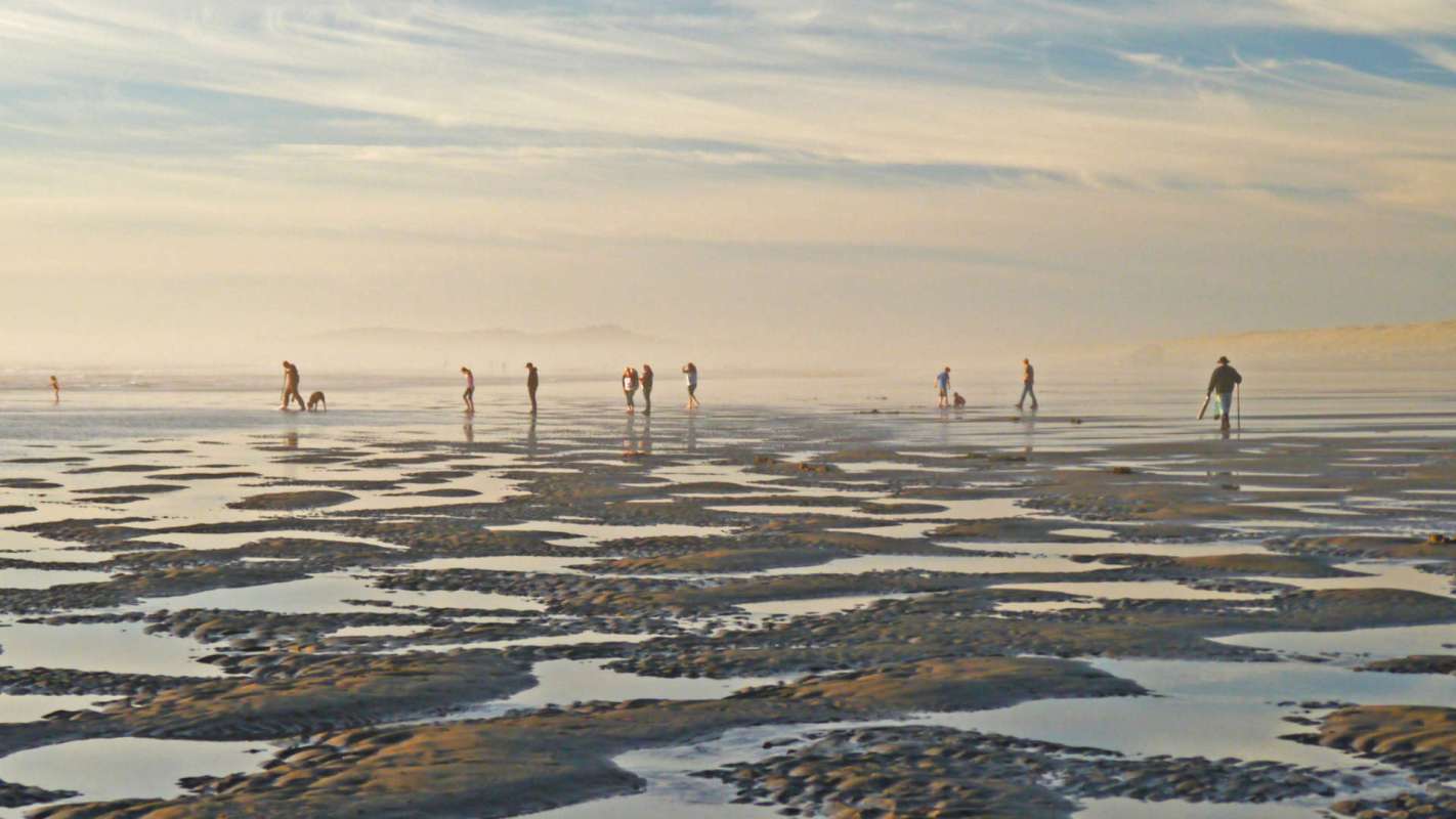 People walking on a beach with shallow water and tidal patterns, under a cloudy sky.