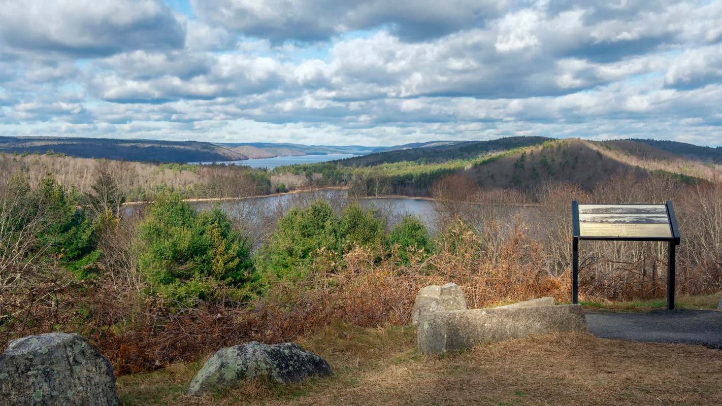 A panoramic view of a lake surrounded by hills and trees, with a signpost in the foreground.
