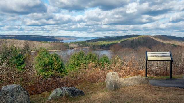 A panoramic view of a lake surrounded by hills and trees, with a signpost in the foreground.