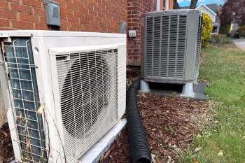 An air conditioning unit and a heat pump next to a brick wall, with mulch and a drainage hose visible on the ground.