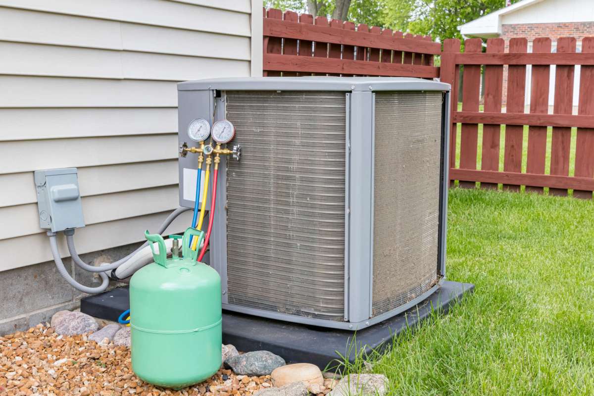 An outdoor air conditioning unit with gauges and a green gas tank beside it, set against a grassy area and wooden fence.