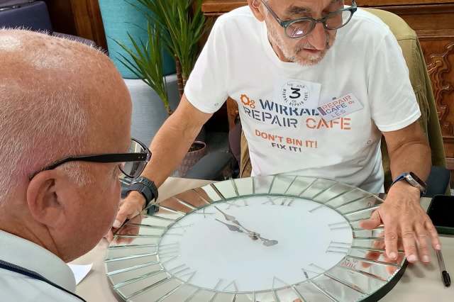 Two men examine a broken clock on a table in a repair café setting.