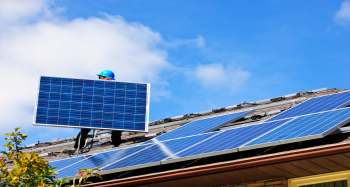 A worker carries a solar panel on a roof with multiple solar panels under a blue sky.