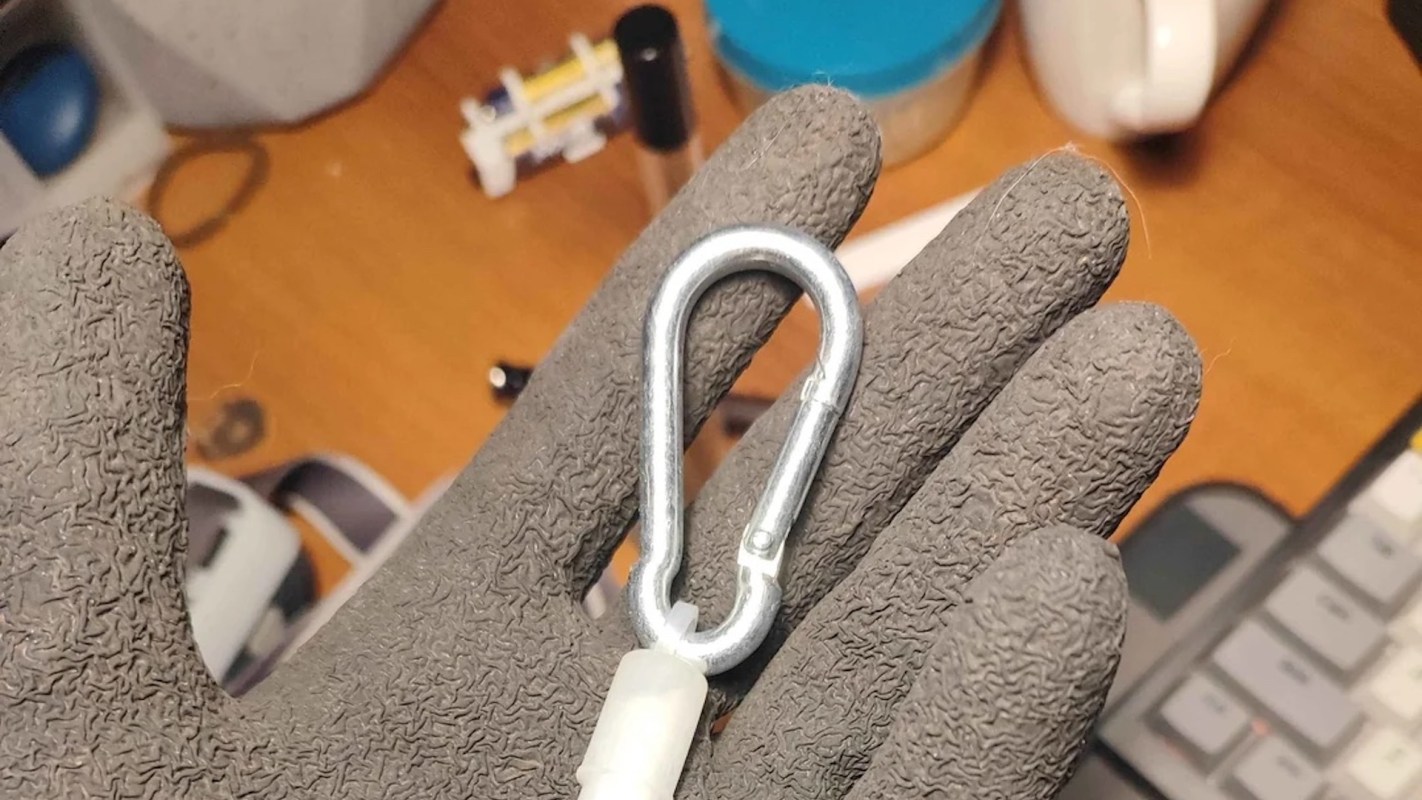 A hand in a glove holds a metal carabiner on a wooden surface with various objects in the background.
