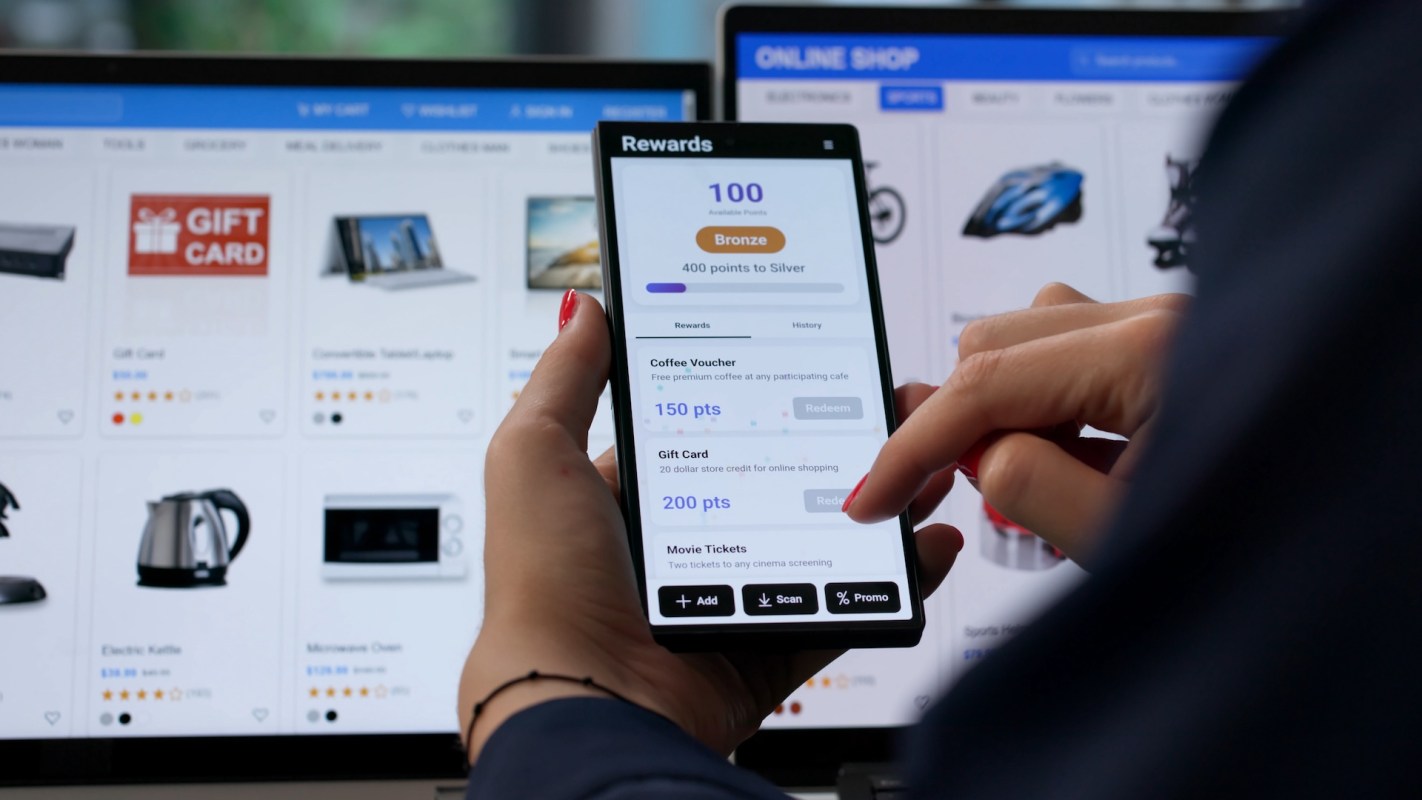 A person interacts with a rewards app on a smartphone with an online shopping website in the background.