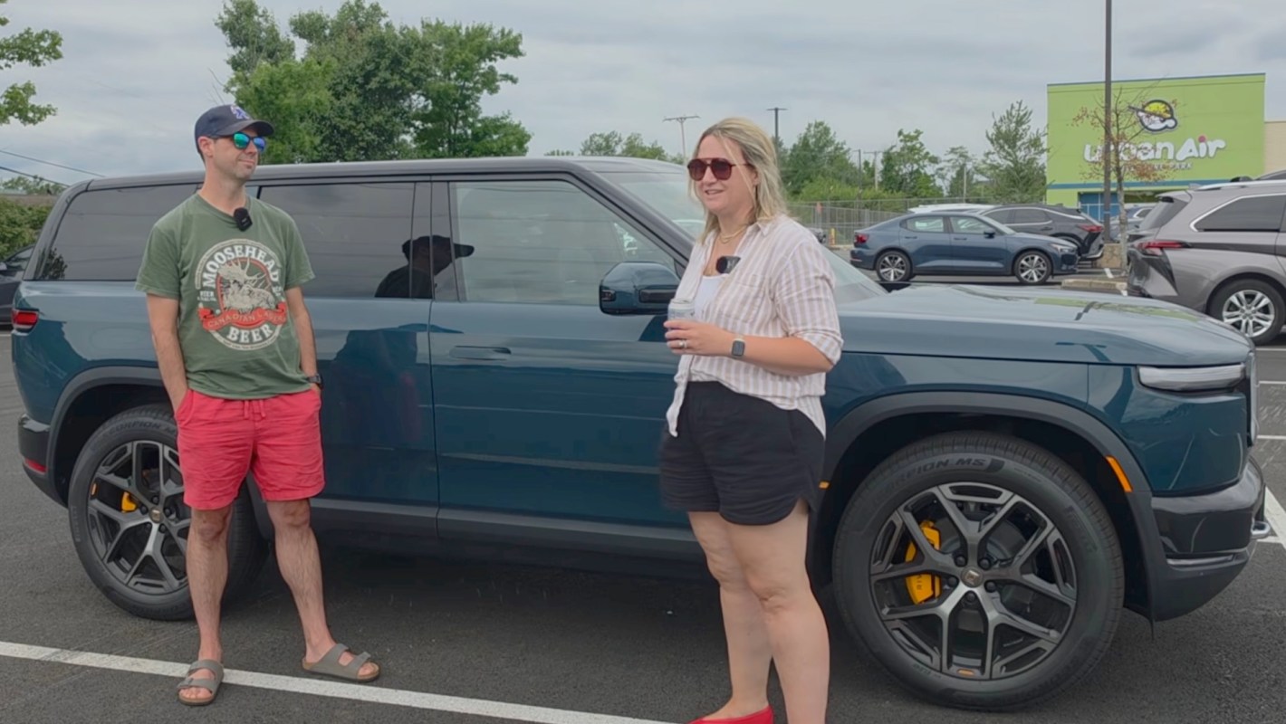 A man and a woman stand beside a blue Rivian SUV in a parking lot.