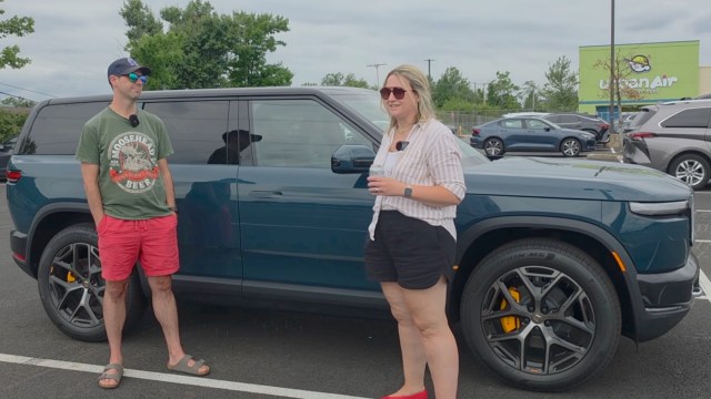 A man and a woman stand beside a blue Rivian SUV in a parking lot.