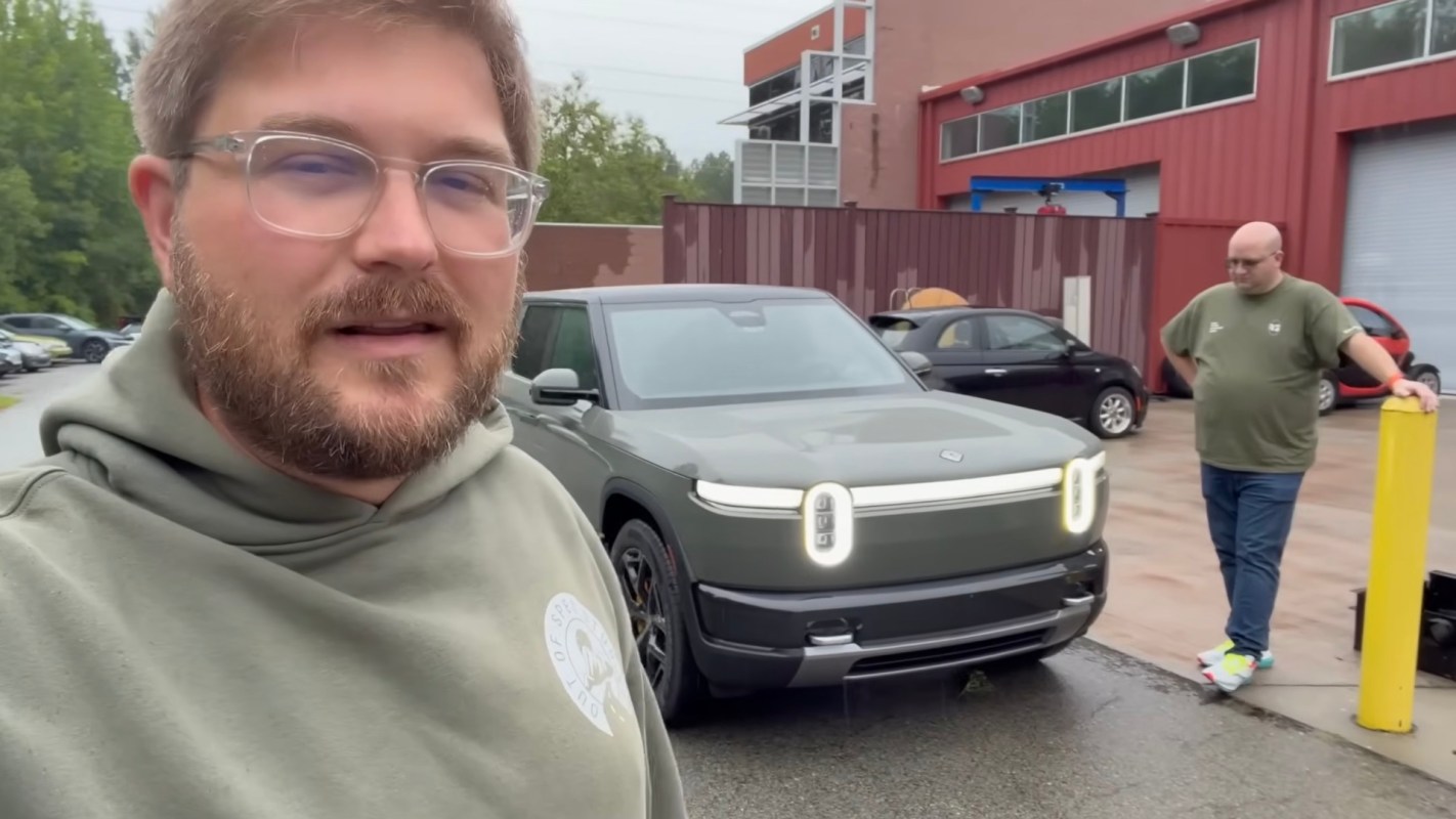 A man stands next to an electric vehicle in a parking lot while another person leans against a post.