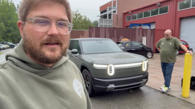 A man stands next to an electric vehicle in a parking lot while another person leans against a post.