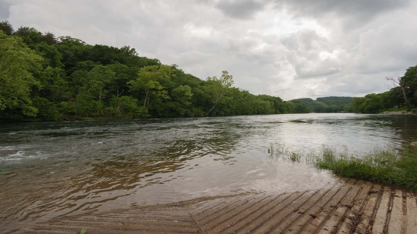 A riverbank bordered by lush green trees under a cloudy sky.