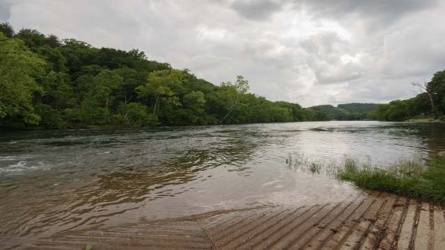 A riverbank bordered by lush green trees under a cloudy sky.