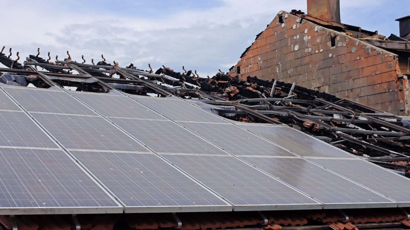Damaged solar panels on a roof with charred remains of a building structure.